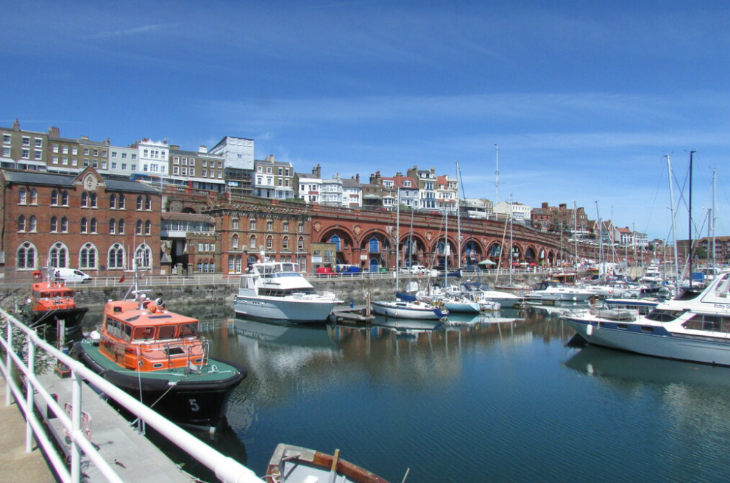 Kent's best seaside towns: A sunny harbor filled with various boats and yachts, set against a backdrop of historic brick buildings and a long arched viaduct under a clear blue sky.