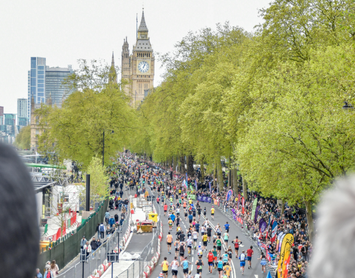 Top things to do in London this weekend: High-angle view of the London Marathon, showing a long road filled with runners and lined with cheering crowds. In the background, the Elizabeth Tower (Big Ben) and the Houses of Parliament rise above a canopy of green spring trees under a bright, overcast sky. The foreground is framed by the out-of-focus heads of two spectators.