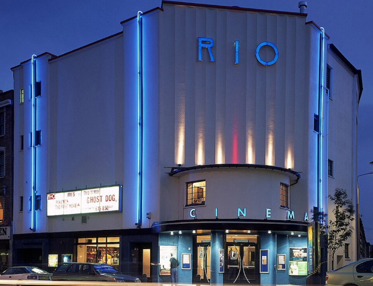 Top events London this weekend: The Art Deco facade of the Rio Cinema in London at dusk, illuminated by blue neon vertical stripes and glowing blue "RIO" and "CINEMA" signs. The white building features a curved entrance with movie posters and a marquee listing "Pokemon The First Movie" and "Ghost Dog."