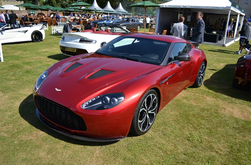 Top events in London this weekend: A metallic red Aston Martin V12 Zagato coupe parked on a grass field at an outdoor car show, with other luxury vehicles and event tents in the background.
