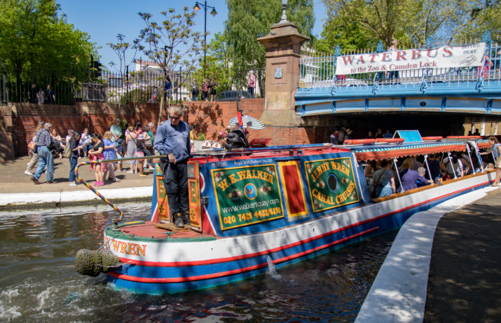 A colorful canal boat named Jenny Wren travels along a waterway in London, passing under a blue bridge with a Waterbus sign, while people watch from the paved bank.