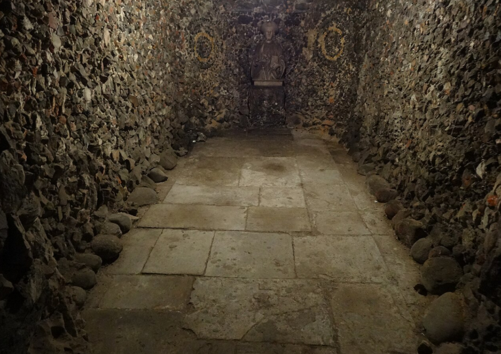 Top events in London today: A dimly lit, narrow stone chamber with walls made of rough, dark volcanic rock and a floor of large, flat paving stones. At the far end, a small stone bust of a man sits on a pedestal against the back wall.