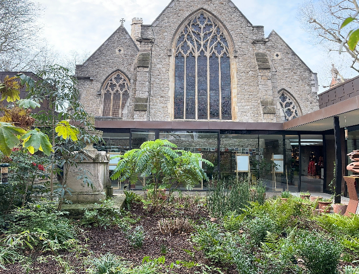 Top events London this weekend: A stone Gothic church with large arched stained-glass windows rises behind a modern glass-walled extension and a lush garden courtyard filled with ferns and green plants.