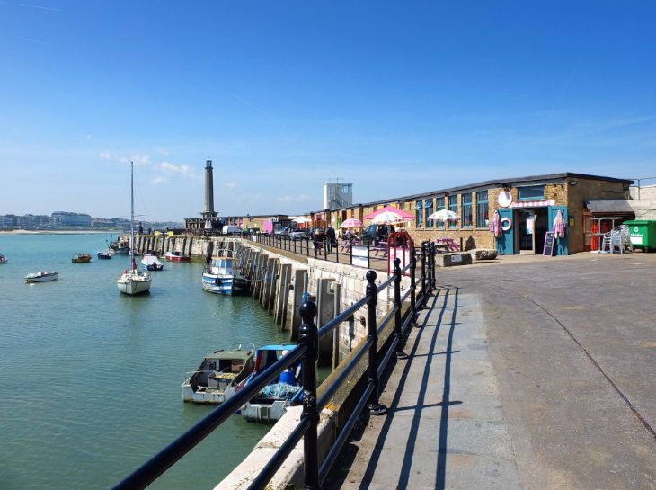 Visit Margate from London day trip: A sunny harbor scene featuring several small boats moored in calm blue water alongside a stone pier. A black metal railing runs along the walkway, which leads past a low brick building with colorful umbrellas and a tall stone lighthouse in the distance.