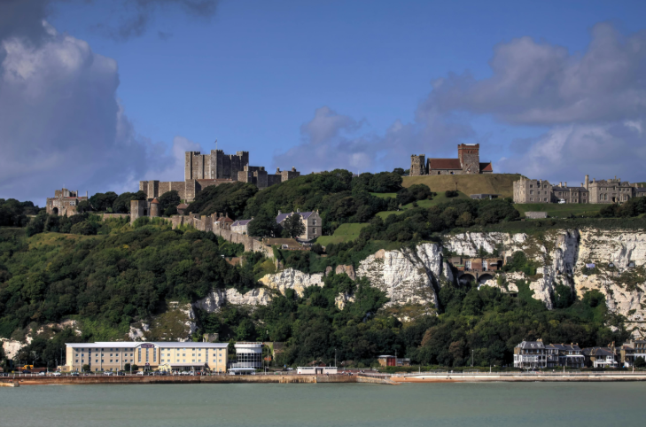 Best seaside day trips in Kent: Dover Castle and St Mary in Castro church sit atop the White Cliffs of Dover, overlooking a coastal town and the sea under a blue sky with scattered clouds.
