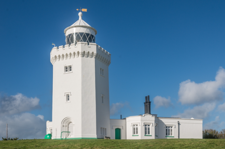 A white, octagonal lighthouse with a black-latticed lantern room and weather vane stands on a grassy hill against a clear blue sky. A small, one-story white building with green doors and window trim is attached to the base.