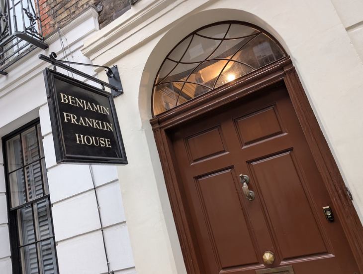 Fourth July/Independence Day 2026 in London:  The entrance to Benjamin Franklin House in London, featuring a dark brown wooden door with a brass knocker and a black hanging sign with gold lettering.