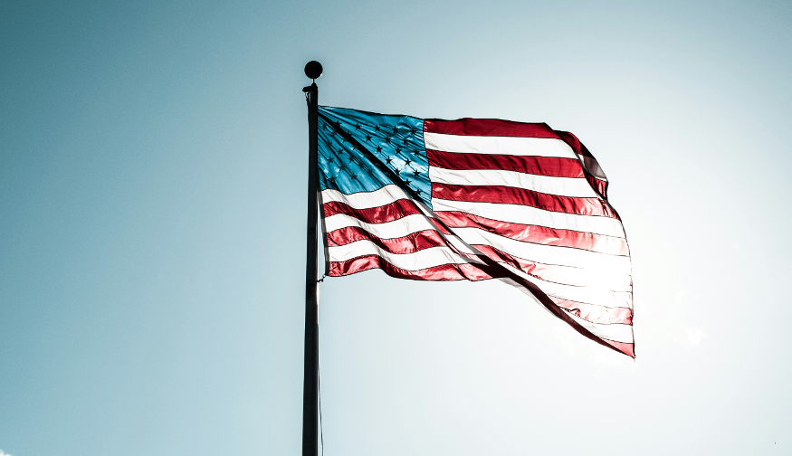 Fourth July/Independence Day 2026 in London:  The American flag waving from a tall flagpole against a bright, clear blue sky with the sun shining through the fabric.