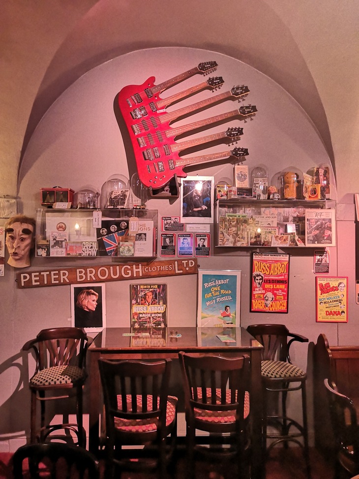 A wall in a pub or cafe decorated with eclectic memorabilia, featuring a large red six-neck electric guitar mounted above a table with chairs. The wall is filled with vintage posters, framed photos, and glass display cases containing various trinkets and collectibles.