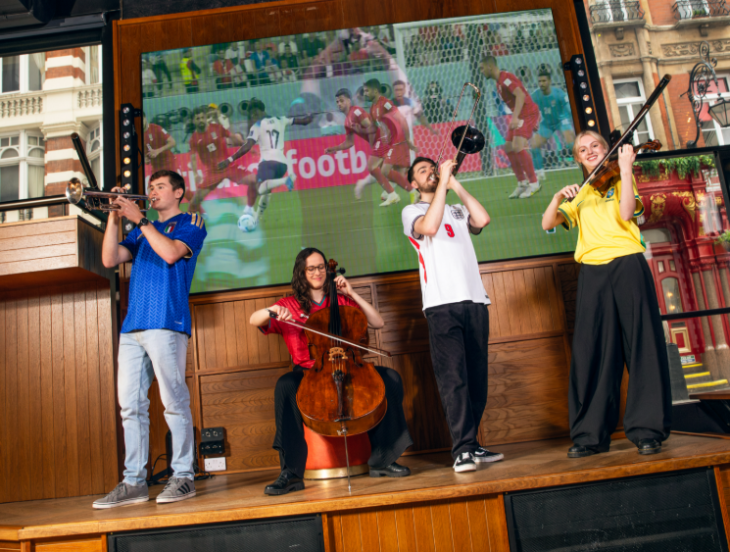 A quartet of musicians on a stage in front of a large screen showing a football match