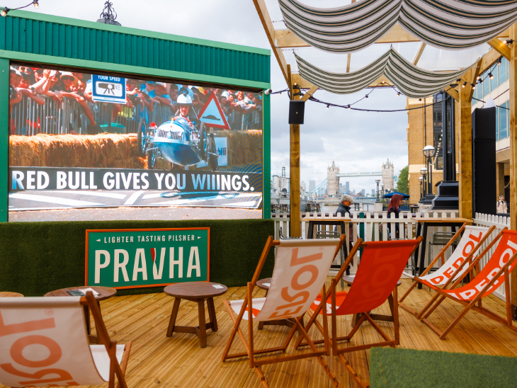 Free screenings of Wimbledon 2026 matches in London: a wooden deck/gazebo with Aperol branded deckchairs facing towards a screen. Tower Bridge is visible in the background