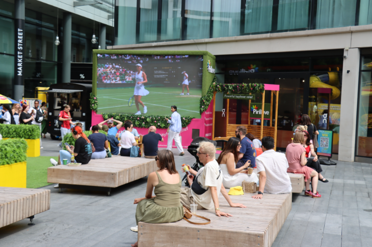Free screenings of Wimbledon 2026 matches in London: people sitting on benches in the open-air in front of a large screen which is showing a tennis match
