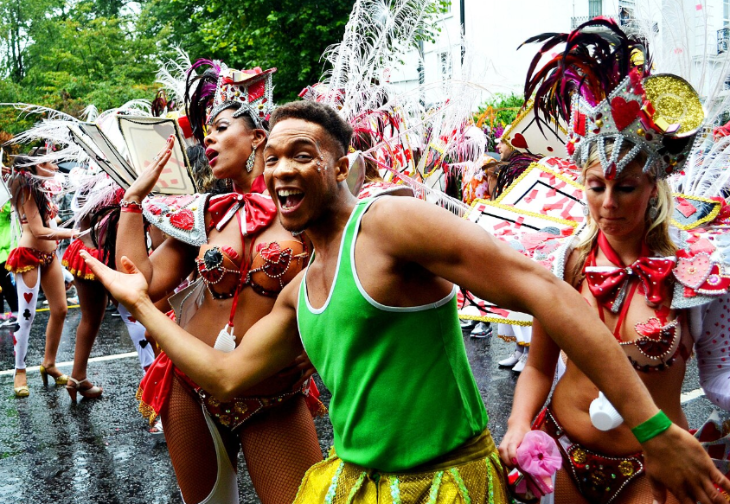 Notting Hill Carnival 2026: A group of people in vibrant, feathered carnival costumes dance in a parade on a wet street. A man in a green tank top and yellow sequined skirt smiles broadly in the foreground, flanked by two women in elaborate red and white outfits with tall, feathered headpieces.