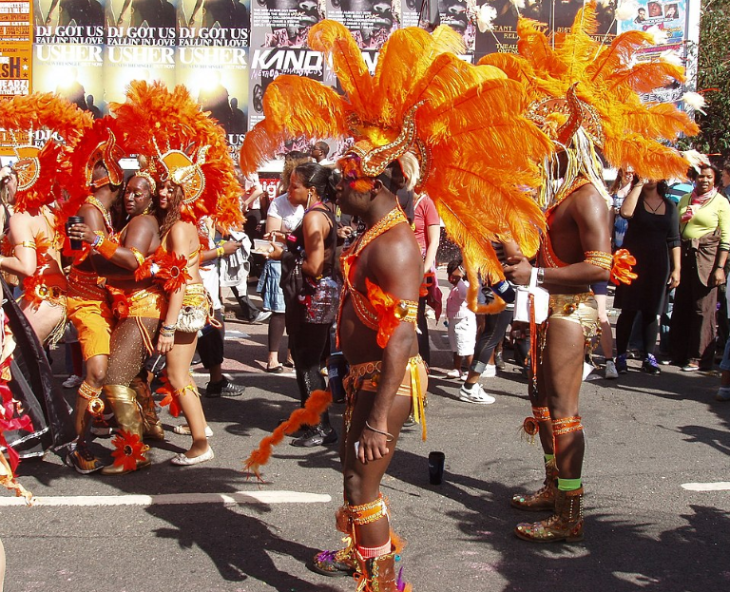 Notting Hill Carnival 2026: A group of carnival performers in vibrant orange feathered headdresses and matching costumes parade down a street lined with posters.
