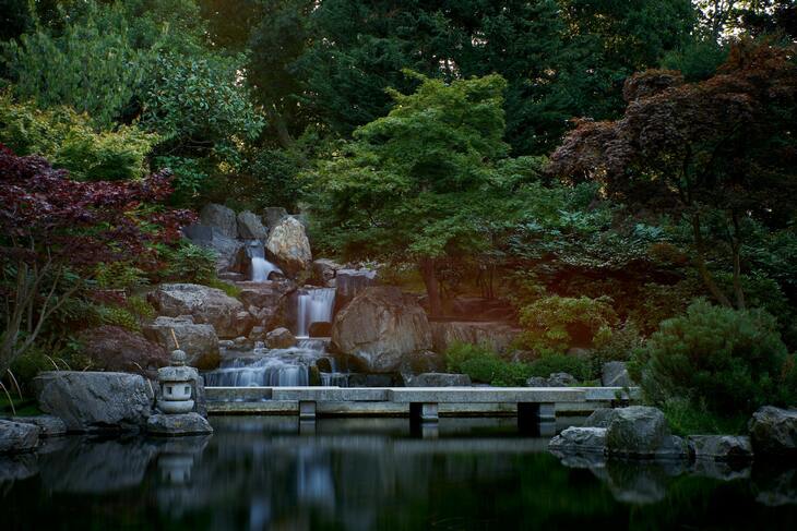 A serene Japanese garden featuring a multi-tiered waterfall flowing over large rocks into a calm pond, crossed by a flat stone bridge and surrounded by lush green and burgundy trees.
