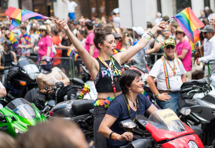 A woman stands on the back of a motorcycle during a Pride parade, holding two rainbow flags high with her arms outstretched. She wears a black crop top with "PRIDE" on it and has a yellow whistle in her mouth. A woman in a blue shirt sits in front of her on the red motorcycle, and other motorcyclists and parade participants are visible in the background.