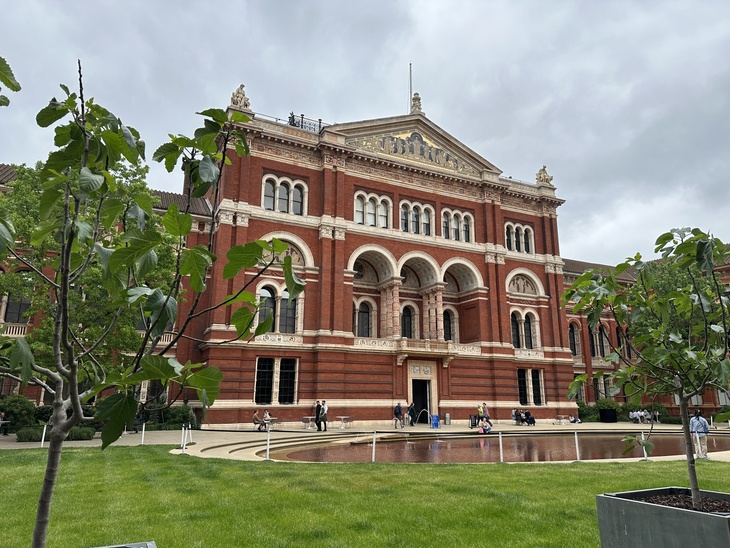 The red brick and terracotta facade of the Victoria and Albert Museum's courtyard in London, featuring ornate arches, a central pediment, and a shallow reflecting pool in the foreground surrounded by a green lawn and small trees.