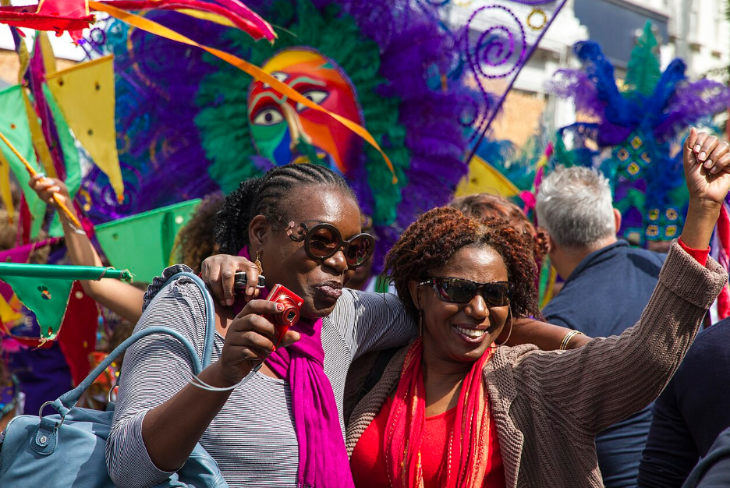 Notting Hill Carnival 2026: Two women smile and embrace at a vibrant carnival, one holding a red camera and the other raising a fist, with colourful costumes and flags in the background.
