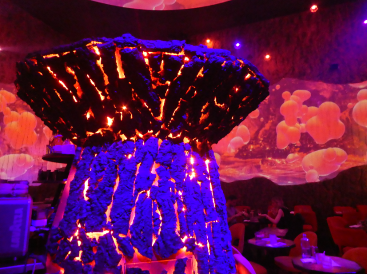Extreme day trip from London to Lille: people sitting at dinner tables in a dimly lit restaurant with magma projections on the walls, and an illuminated core at the centre