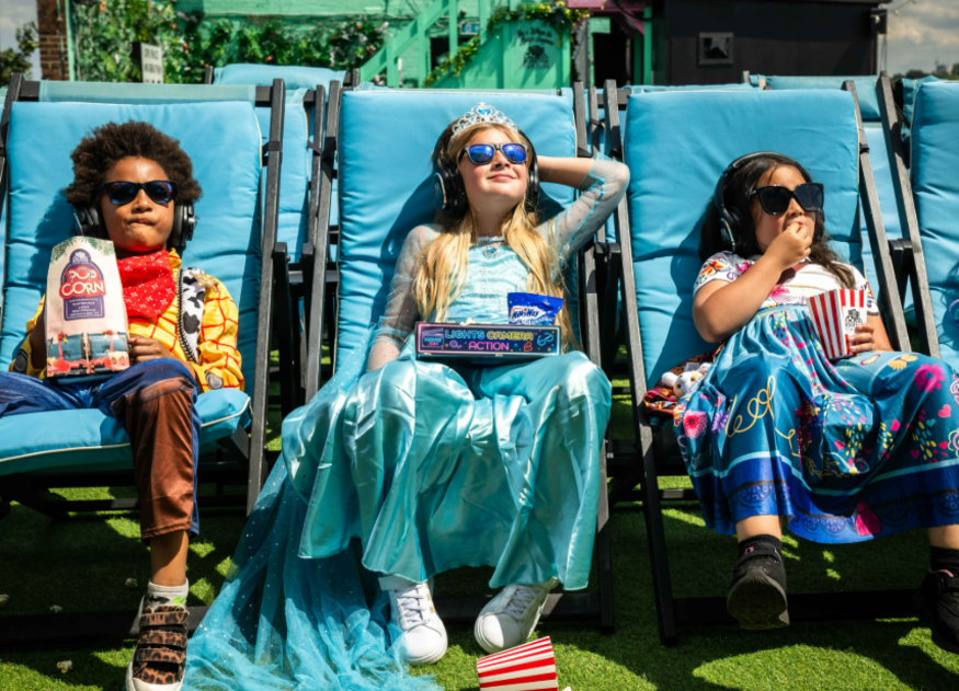 Things to do in London in the summer holidays 2026: three children in fancy dress costumes sitting in deckchairs in the sun