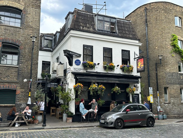 Fourth July ceelbrations in London 2026: A historic white-walled pub called The Mayflower on a cobbled London street, featuring black window frames, flower boxes, and outdoor seating with patrons. A grey Fiat 500 is parked in front.
