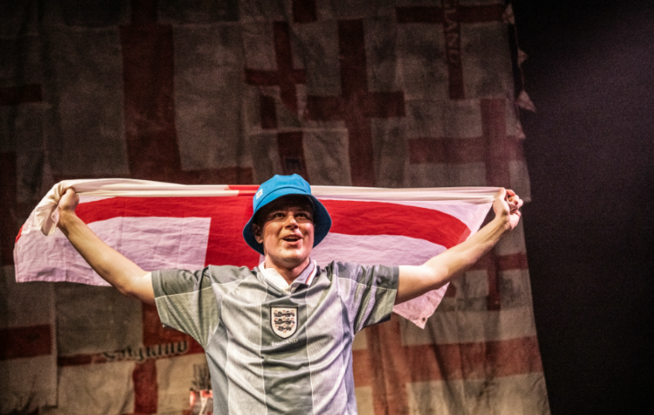 What's on in London in June 2026: an actor on stage wearing an England football shirt and bucket hat, holding the England flag up behind him