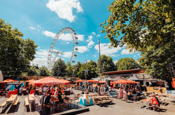 Top bank holiday weekend events London: people sitting at picnic benches in the sunshine, with the London Eye in the bakcground