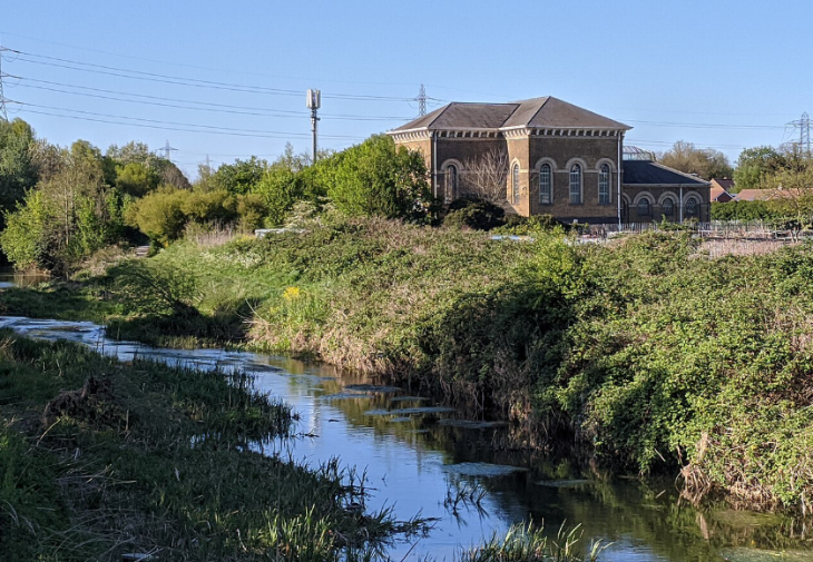 Things to do in London this weekend: A brick building with arched windows stands on a grassy bank overlooking a narrow, winding river under a clear blue sky. Power lines and a cell tower are visible in the background.