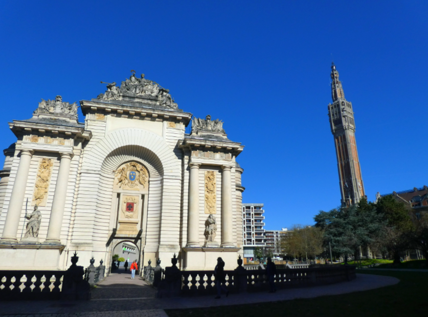 Extreme day trip from London to Lille: a structure like the Arc de Triomphe in the foreground, and a tall redbrick tower in the background, beneath a blue sky