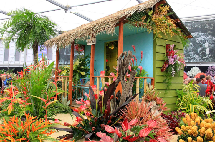 Top bank holiday weekend events London: A tropical garden display featuring a small wooden shack with a thatched roof and light blue interior. The shack is surrounded by lush greenery, palm trees, and vibrant exotic flowers in shades of red, orange, and pink. People are visible in the background of the indoor exhibition space.