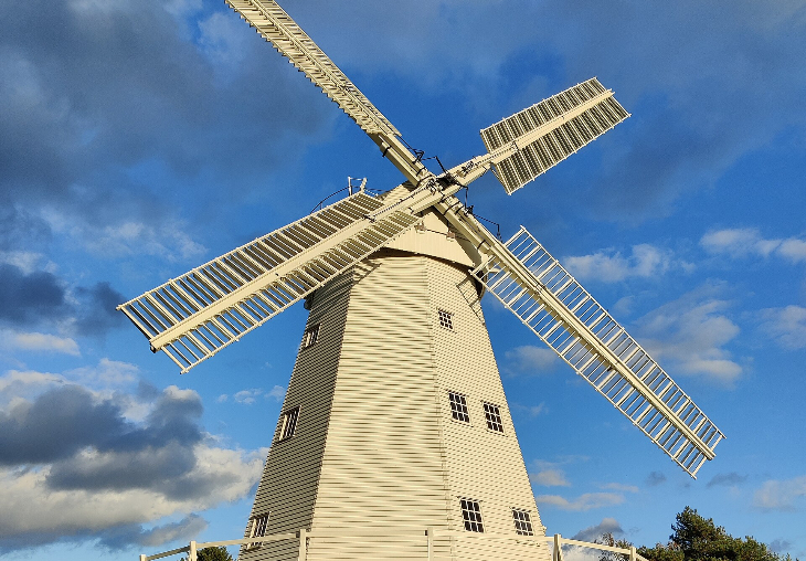 Top bank holiday weekend events London: A white, wooden smock windmill with four large sails stands against a blue sky with scattered clouds. The windmill features a brick base, a wrap-around wooden gallery, and several small windows.