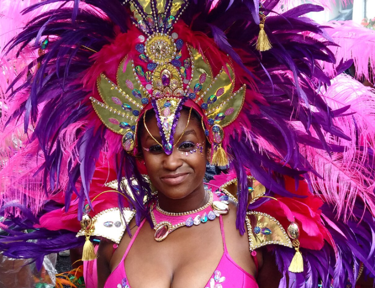 Top things to do in London in summer: A smiling woman in a vibrant pink and purple carnival costume with a large feathered headdress and jeweled accessories.