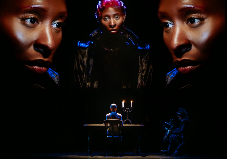 Cynthia Erivo on stage, sitting at a desk with her back to the audience, with three large projections of her face above