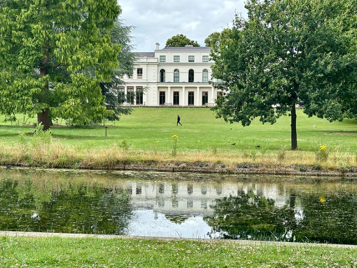 A large, white neoclassical mansion sits atop a grassy hill, reflected in a calm pond in the foreground. A lone person walks across the lawn between the house and the water, framed by large green trees on either side.
