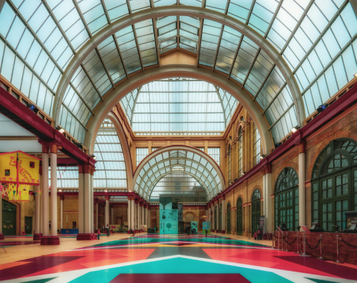 What's on in London today: A wide-angle view of a grand, Victorian-style hall with a high vaulted glass ceiling and arched steel supports. The floor is covered in a vibrant, geometric pattern of pink, teal, and red, leading toward a central exhibit in the distance.