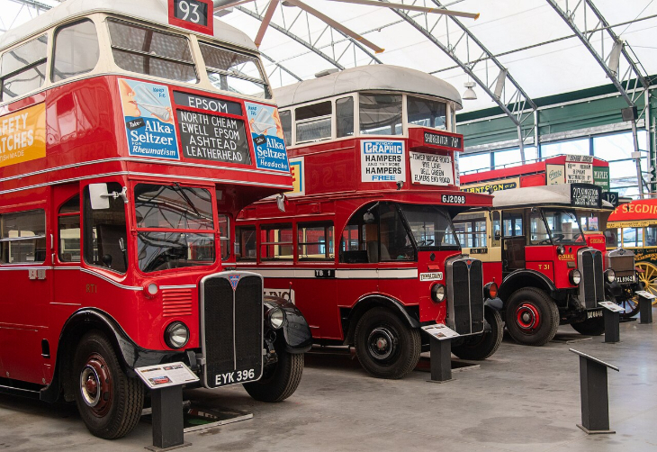 Things to do this weekend: A row of vintage red double-decker and single-decker London buses parked inside a museum hangar with a vaulted glass ceiling. The bus in the foreground features advertisements for Alka-Seltzer and a destination board for Epsom.