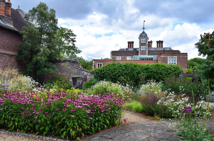 Top weekend events London: A lush garden with vibrant pink coneflowers and white wildflowers in the foreground, leading to a historic brick manor house with a central clock tower under a cloudy sky.