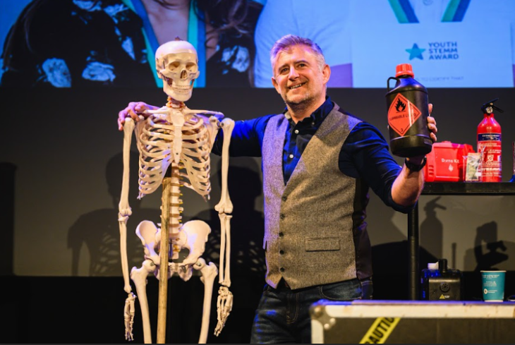 Things to do in London this weekend: a man on stage with one arm around a skeleton, holding a jar marked as flammable in his other hand