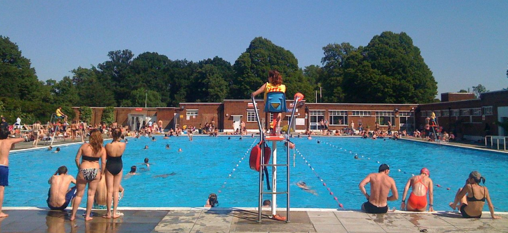 London's best outdoor swimming pools and lidos: A sunny outdoor public swimming pool filled with people, featuring a lifeguard on a high chair in the foreground and brick buildings and trees in the background.