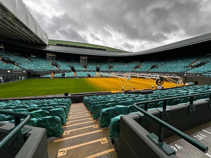 Where to watch Wimbledon 2026 matches: Wide-angle view of an empty tennis stadium under a cloudy sky, with rows of green seats covered in plastic and specialized lighting rigs positioned over the grass court.