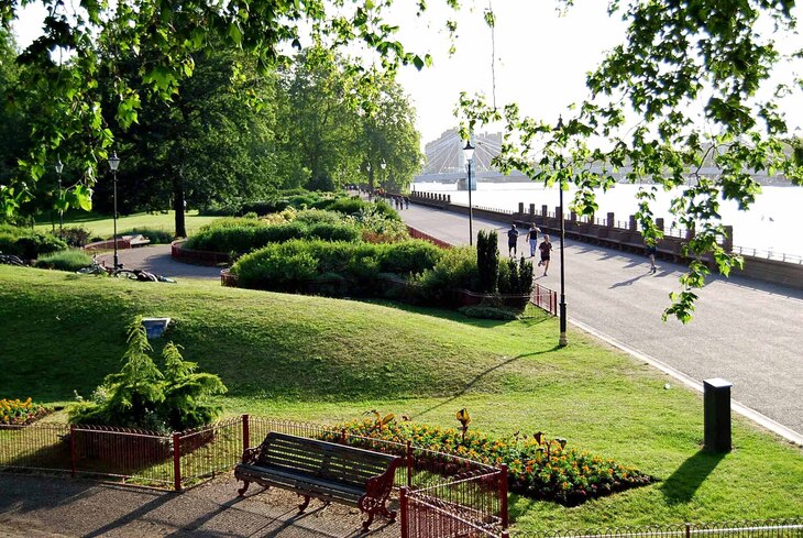 A sunny park scene with a paved path running alongside a river, where people are jogging. In the foreground, there is a wooden bench, flower beds, and lush green lawns framed by leafy trees.