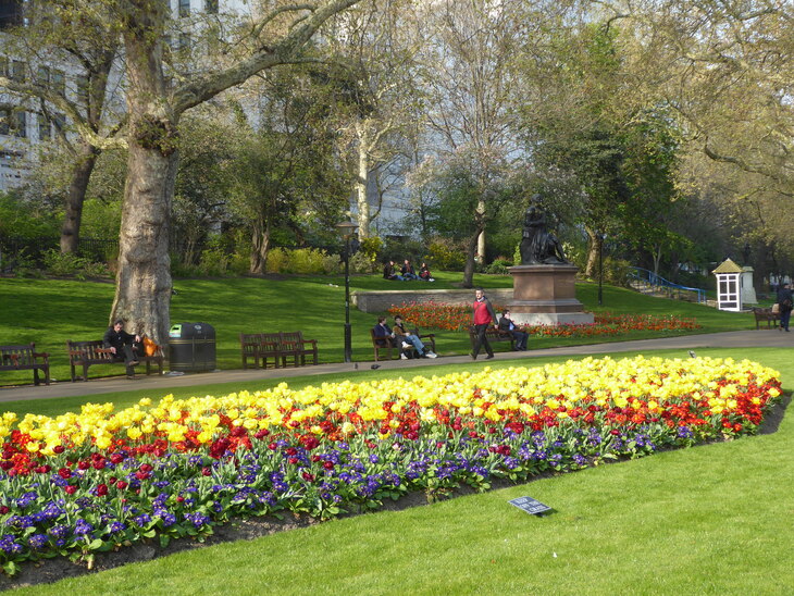 A vibrant public park featuring a long, tiered flowerbed of yellow, red, and purple flowers in the foreground. People sit on benches and walk along a path lined with green lawns, mature trees, and a bronze statue on a stone pedestal.