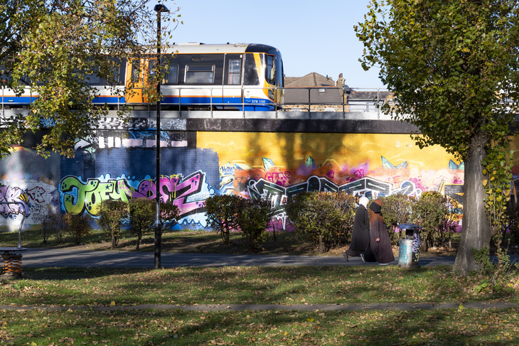 A London Overground train passes on an elevated track above a long wall covered in colorful graffiti. In the foreground, two people in hijabs walk along a path in a park with green grass and autumn leaves.