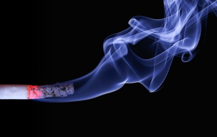 A lit cigarette with a glowing red tip and a long trail of blue smoke swirling against a black background.