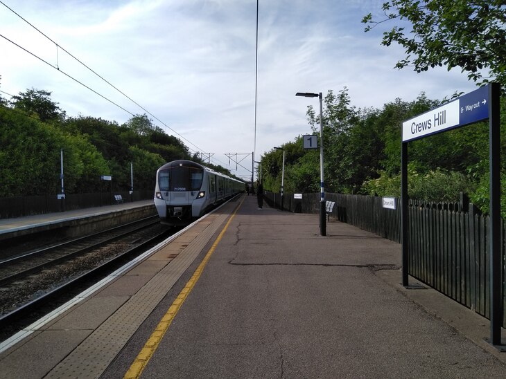 A white and grey train pulls into Crews Hill station platform 1 on a sunny day. A blue and white sign for Crews Hill is in the foreground.