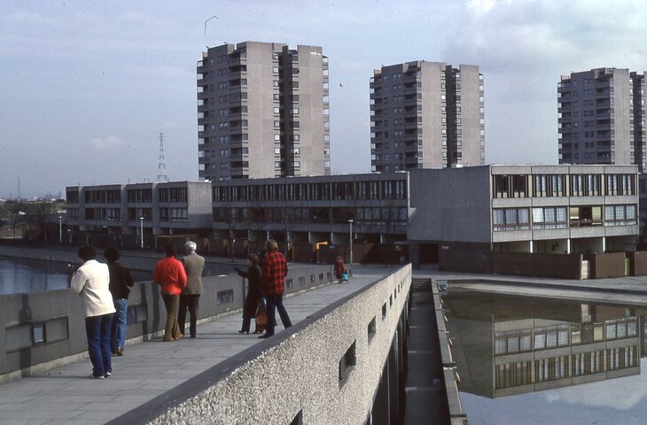 A group of people walk across a concrete pedestrian bridge overlooking a canal and a brutalist housing estate with low-rise blocks and three tall towers.