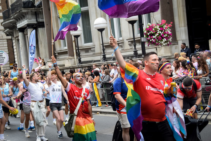 A diverse group of people march in a Pride parade on a city street, waving rainbow and bisexual pride flags. Many participants wear colorful clothing, face paint, and jerseys with "1Password" and "UBS" logos. Spectators and historic buildings line the background.