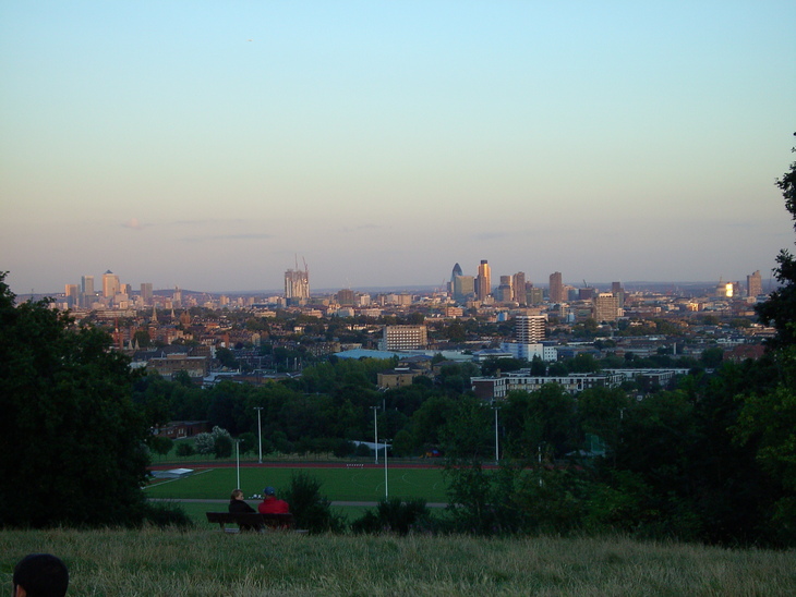 A wide-angle view of the London skyline at dusk from a grassy hill, with a sports field in the foreground and two people sitting on a bench.