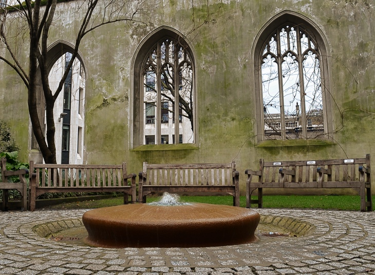 A low-angle shot of a circular stone fountain in a courtyard, with three wooden benches arranged in front of the mossy, windowless stone walls of a ruined Gothic church.