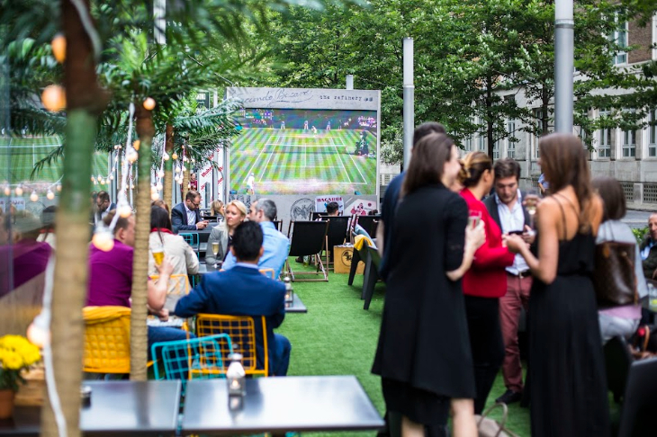 Free screenings of Wimbledon 2026 matches in London: people sitting and standing around in an open-air pub-bar, which has a large screen showing tennis at the far end